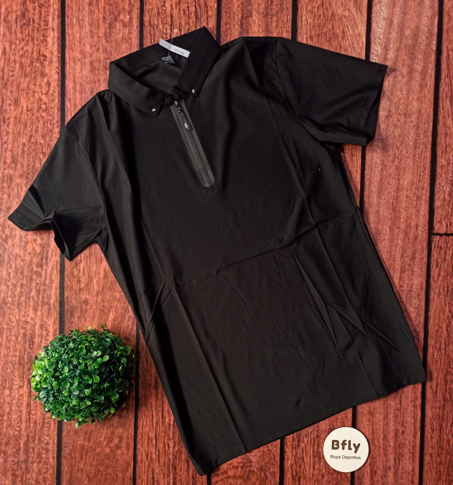 Cuatro camisas abotonadas dobladas en negro, beige, blanco y azul sobre un suelo de madera con una pequeña planta verde.