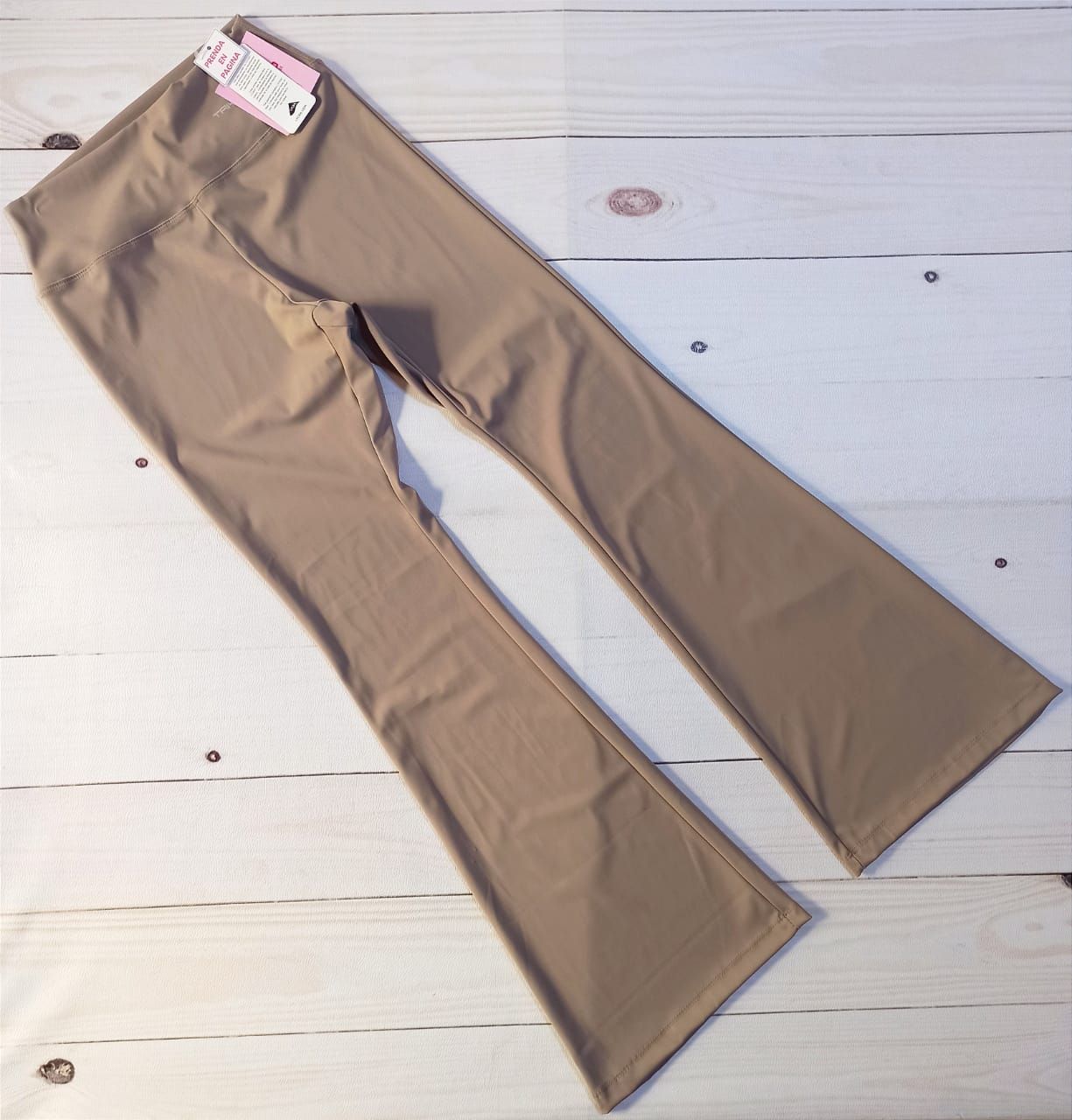 Pantalones acampanados de color caqui tostado extendidos sobre una superficie de madera blanca.