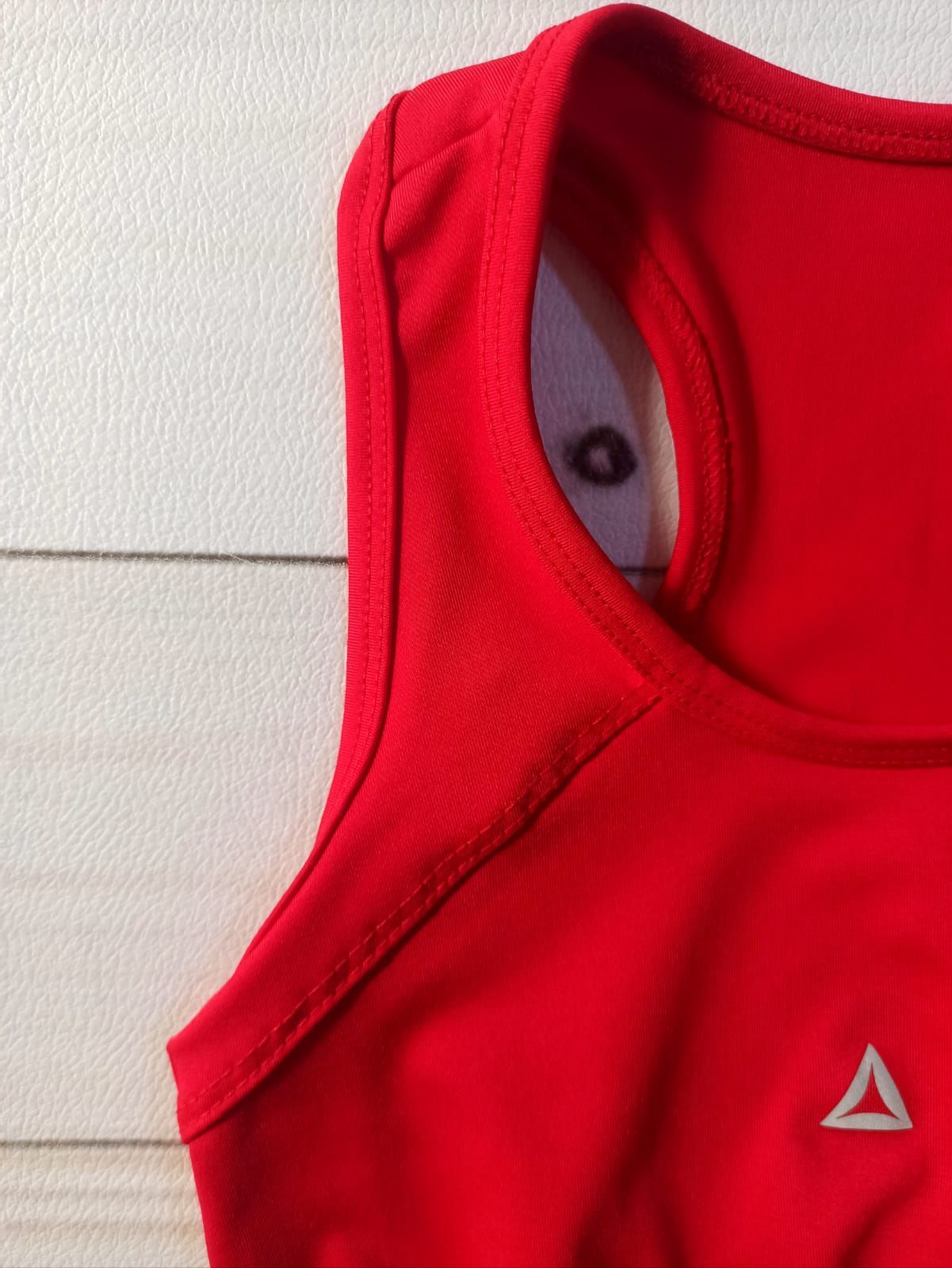 Camiseta sin mangas deportiva corta de color rojo sobre el torso de un maniquí, con un pequeño logotipo triangular en el pecho.