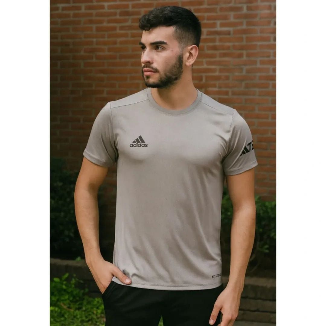 Un hombre, de pie al aire libre, viste una camiseta beige y pantalones negros, y mira de reojo contra una pared de ladrillos.