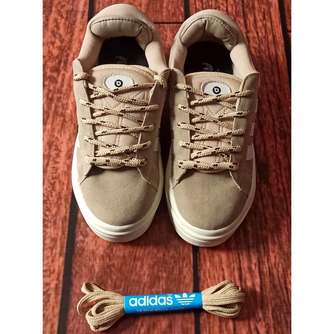 Un par de zapatillas Adidas beige con cordones trenzados sobre un suelo de madera, un paquete de cordones azules al lado.