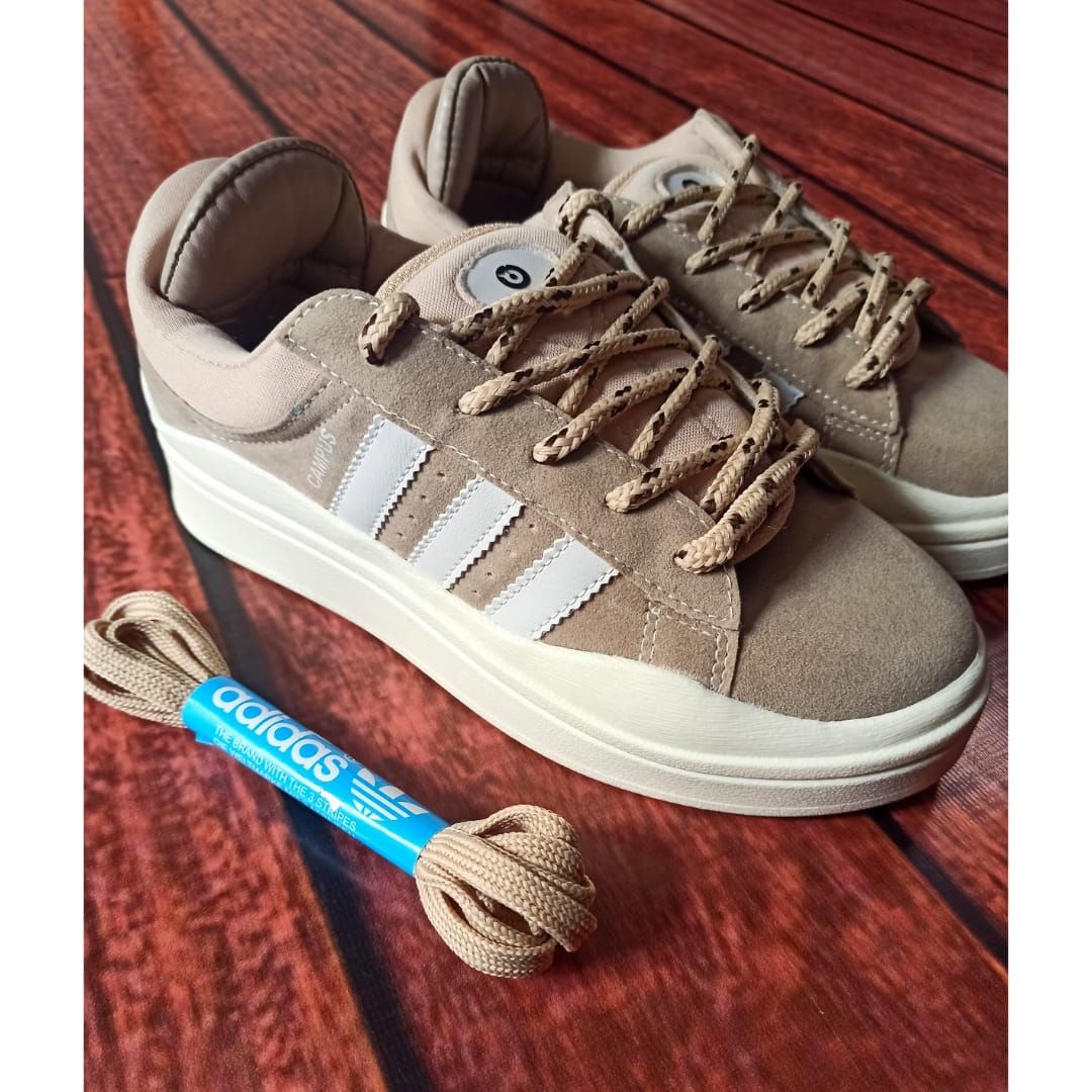 Un par de zapatillas Adidas beige con cordones trenzados sobre un suelo de madera, un paquete de cordones azules al lado.