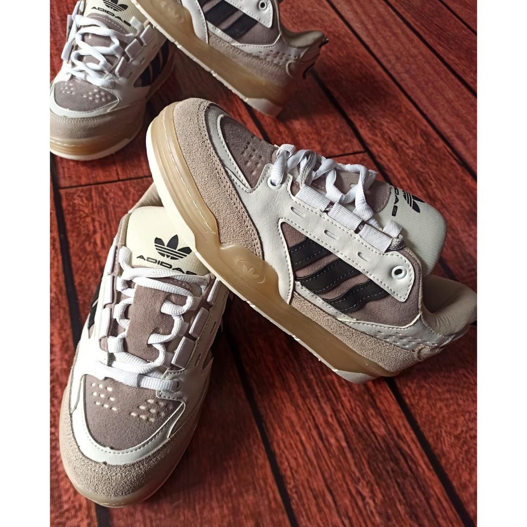 Tres zapatillas Adidas grises y blancas con suelas color canela dispuestas sobre un suelo de madera rojiza.