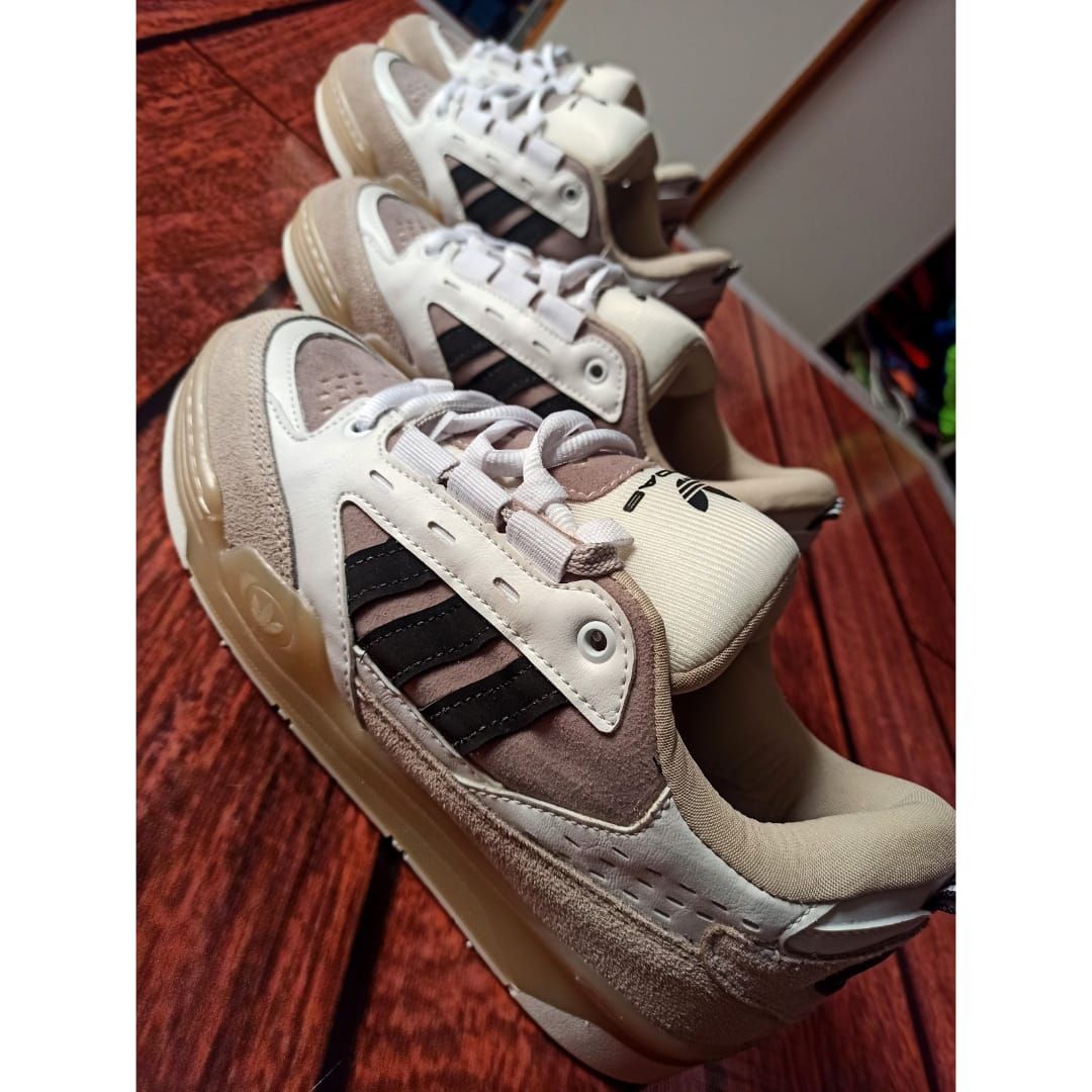 Tres zapatillas Adidas grises y blancas con suelas color canela dispuestas sobre un suelo de madera rojiza.