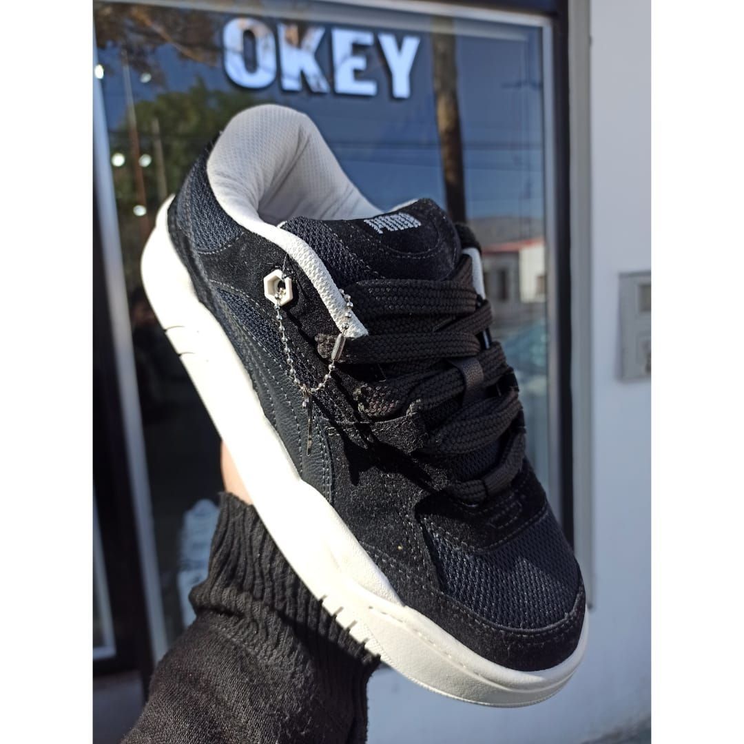 Una zapatilla negra con suela blanca sostenida frente al escaparate de una tienda OKEY.