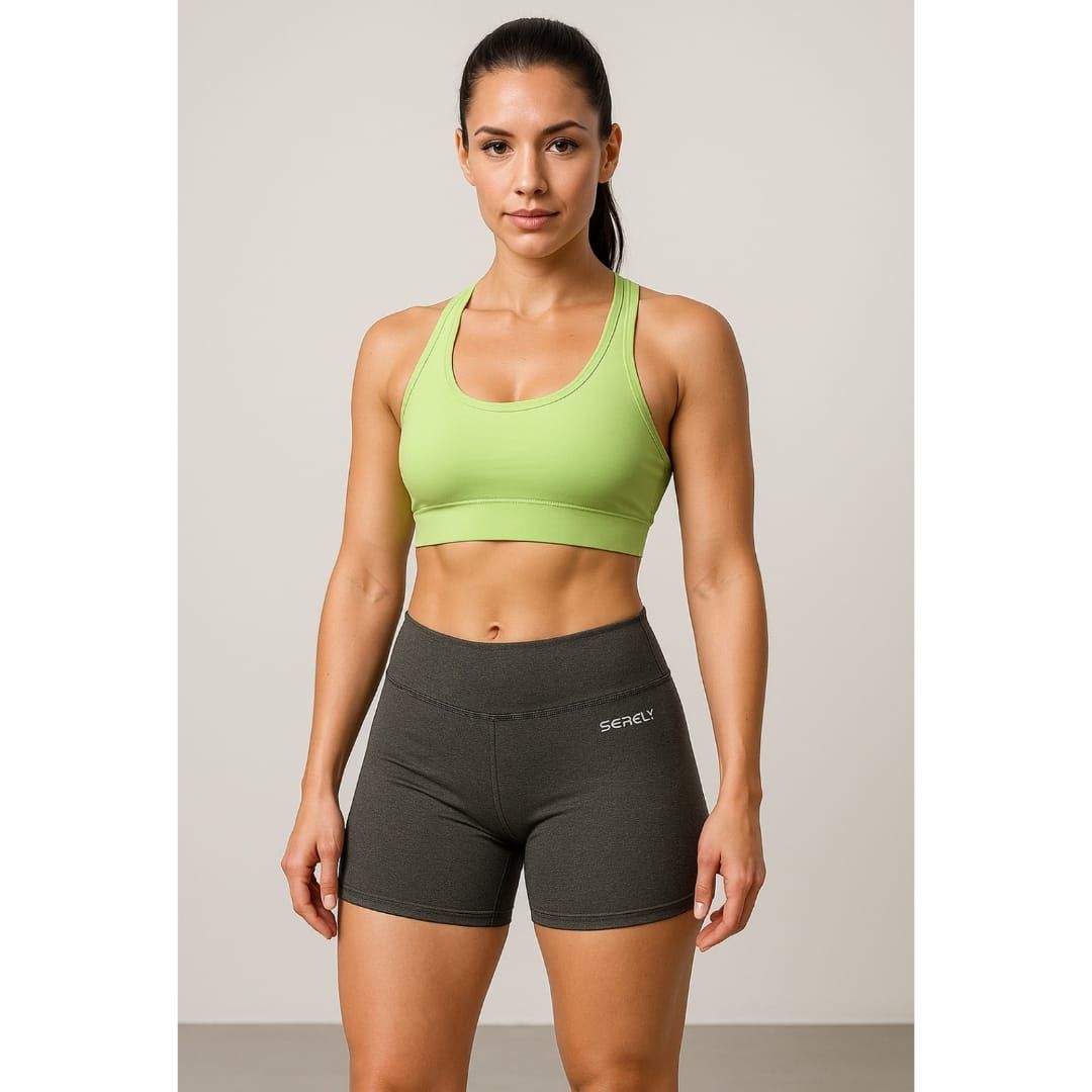 Mujer con sujetador deportivo verde neón y pantalones cortos oscuros de pie frente a un fondo de estudio liso.