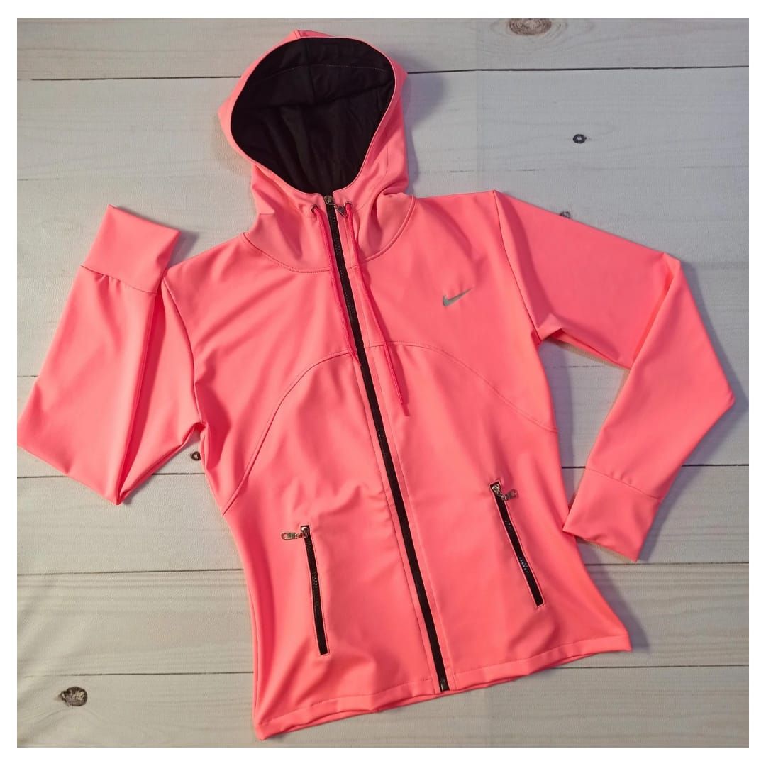 Mujer con chaqueta con capucha y cremallera color coral y pantalones negros sobre un fondo gris liso.