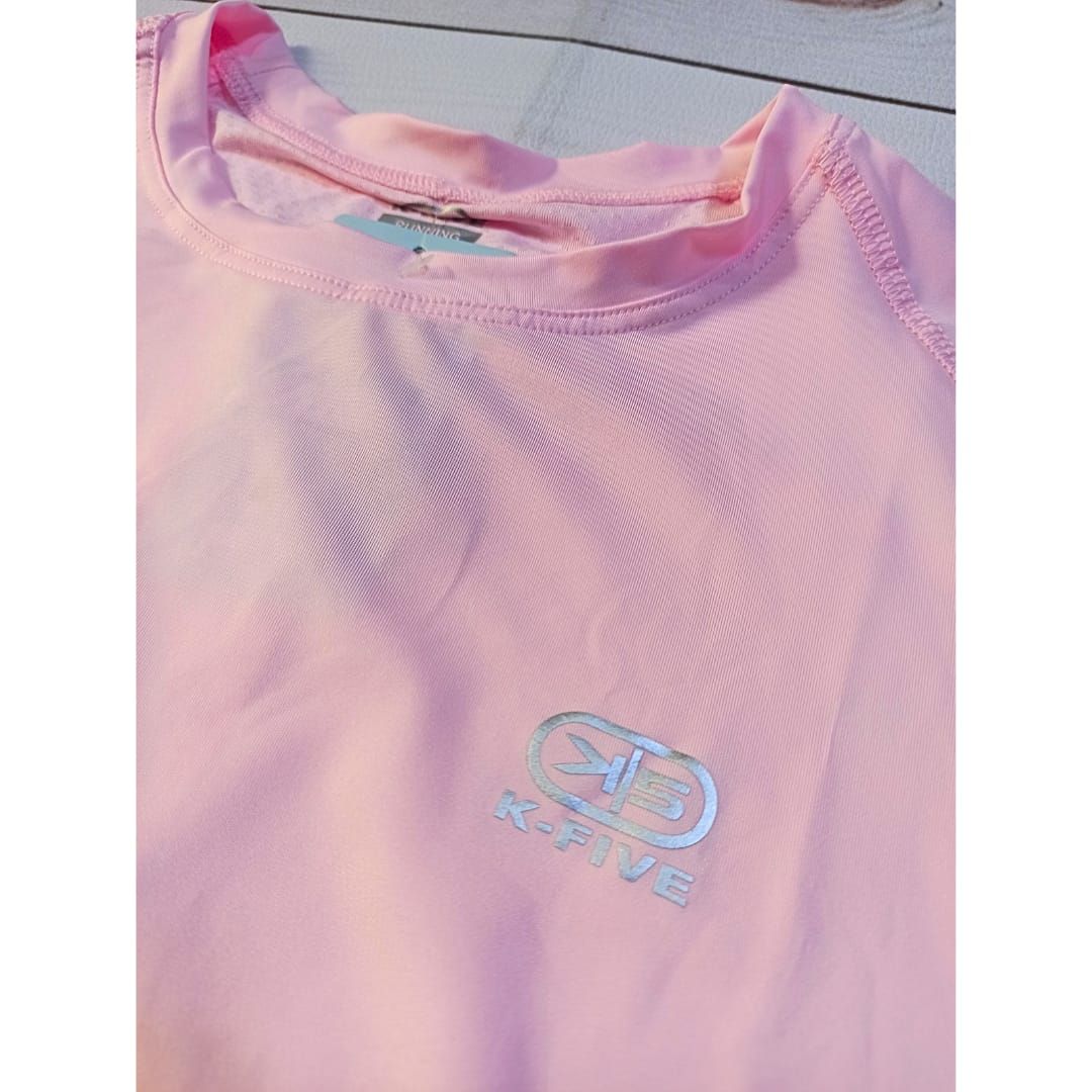 Camisa rosa de manga larga extendida sobre un fondo de madera blanca junto a una pequeña planta verde.