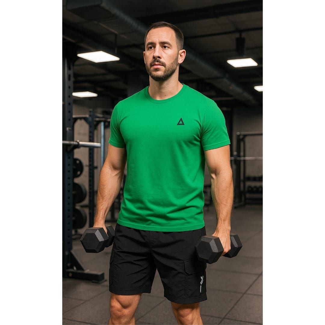 Hombre de pie en un gimnasio sosteniendo mancuernas, vistiendo una camisa verde y pantalones cortos negros.