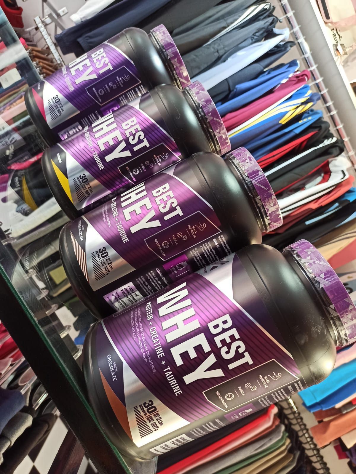 Envase morado de suplemento de proteína de suero en exhibición, con percheros de ropa difuminados al fondo.