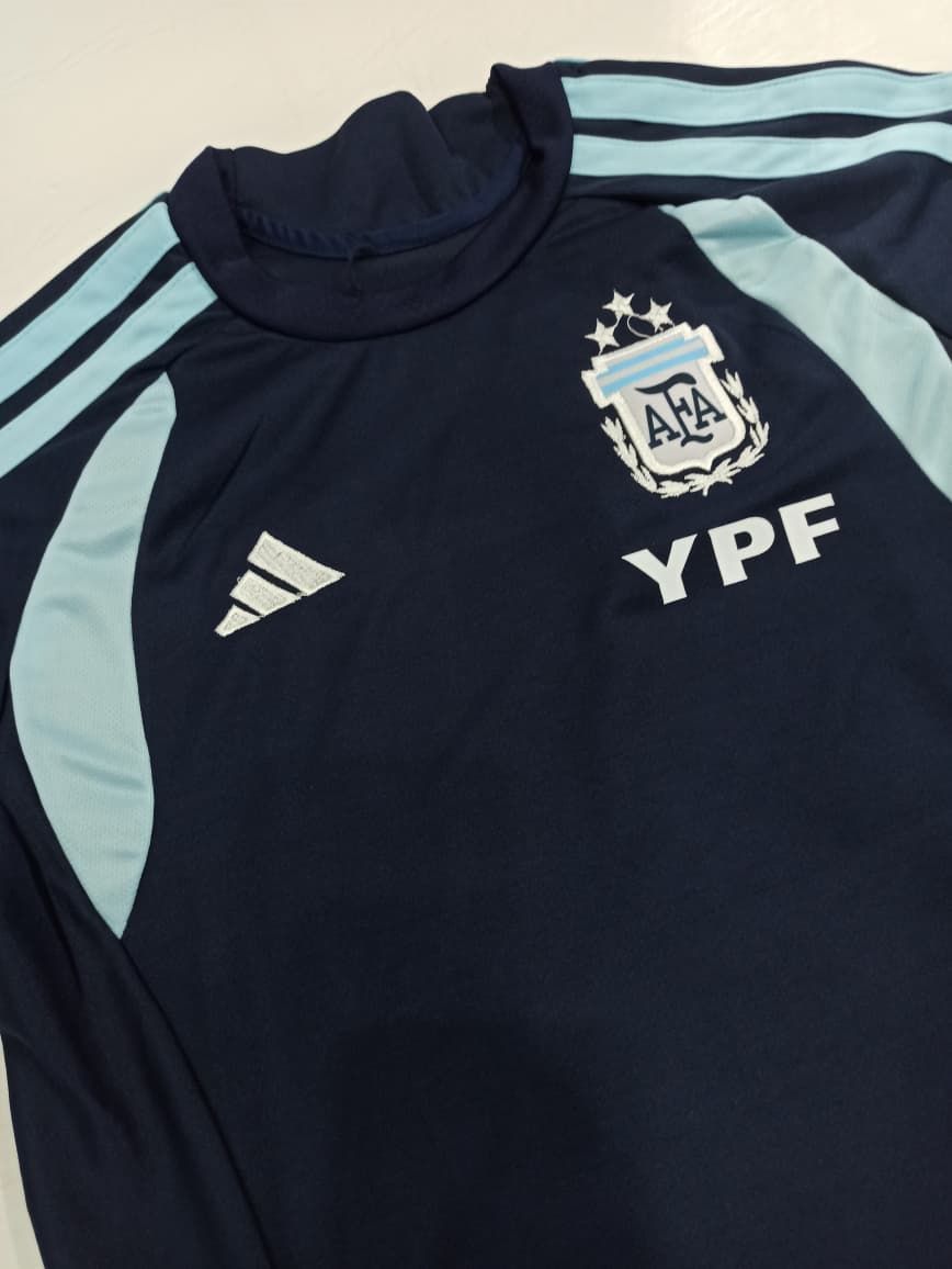 Camiseta de la selección argentina de fútbol, azul marino oscuro con mangas azul claro y el logotipo del patrocinador YPF, colocada sobre una superficie plana.