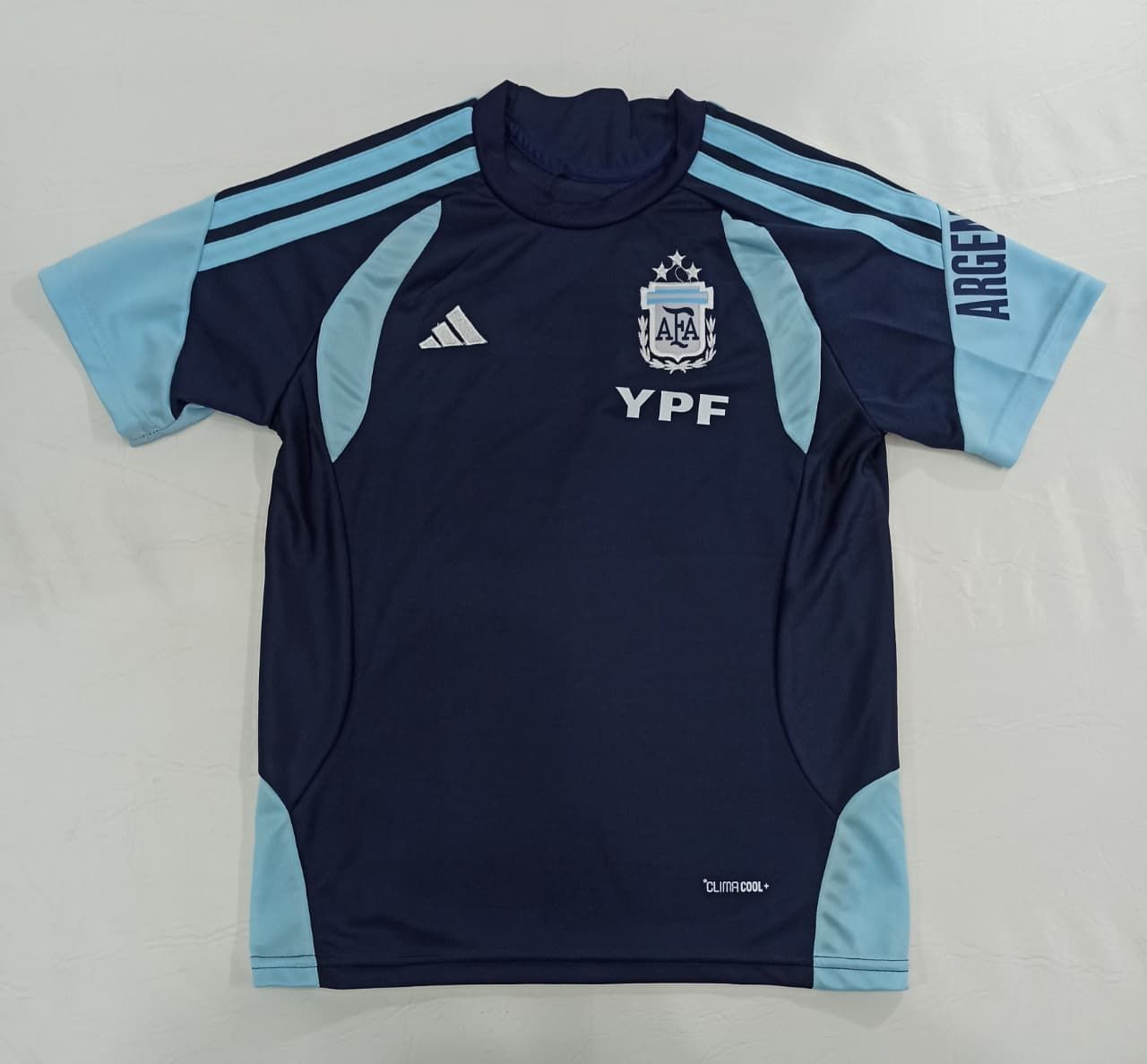 Camiseta de la selección argentina de fútbol, azul marino oscuro con mangas azul claro y el logotipo del patrocinador YPF, colocada sobre una superficie plana.