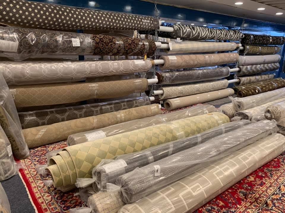 Stacks of rolled carpets and rugs in a showroom, wrapped in clear plastic and arranged on shelves.