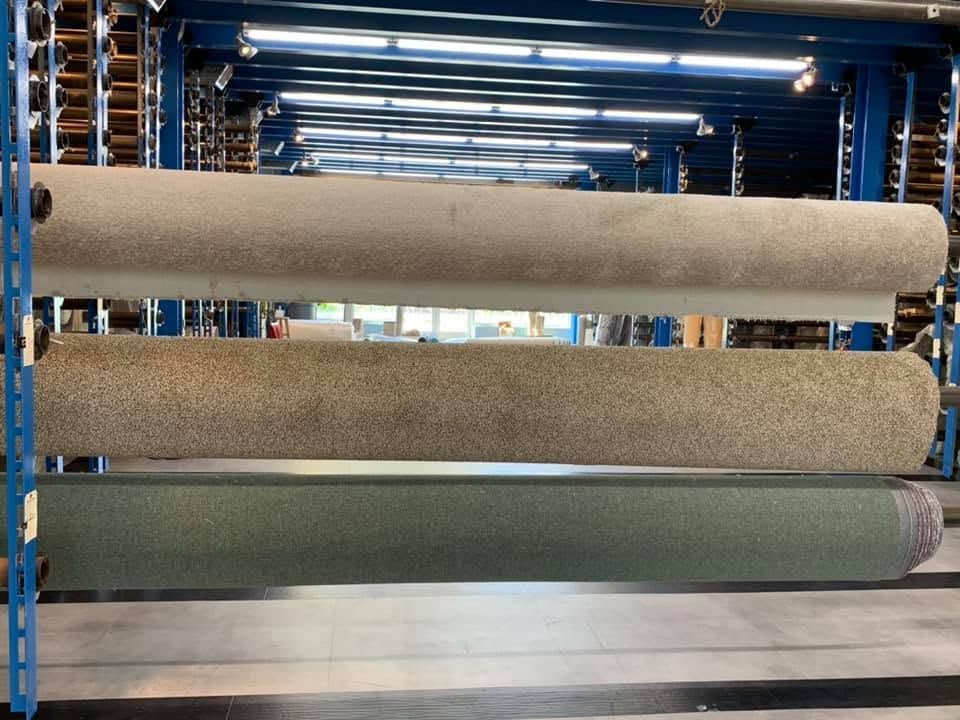 Three large rolls of carpet or fabric stacked on blue industrial racks in a warehouse