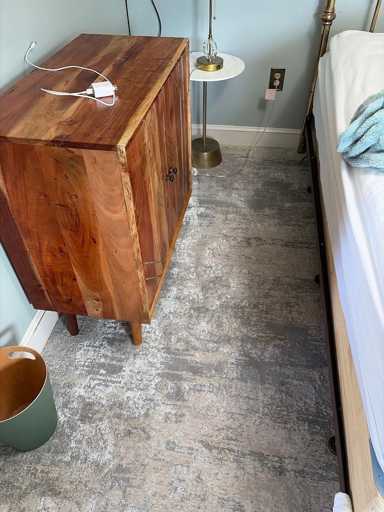 Narrow bedroom corner with wooden dresser, lamp, bed edge, and gray carpet.