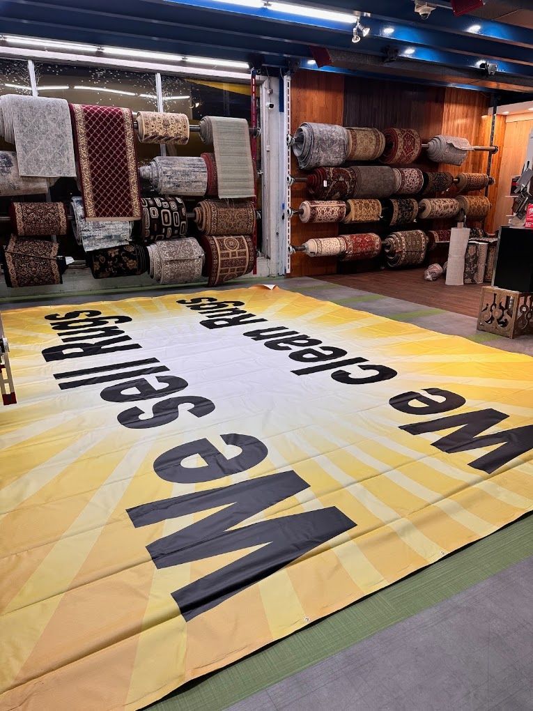 Large “We Sell Rugs” floor banner in a rug warehouse with stacked rolled carpets on shelves.