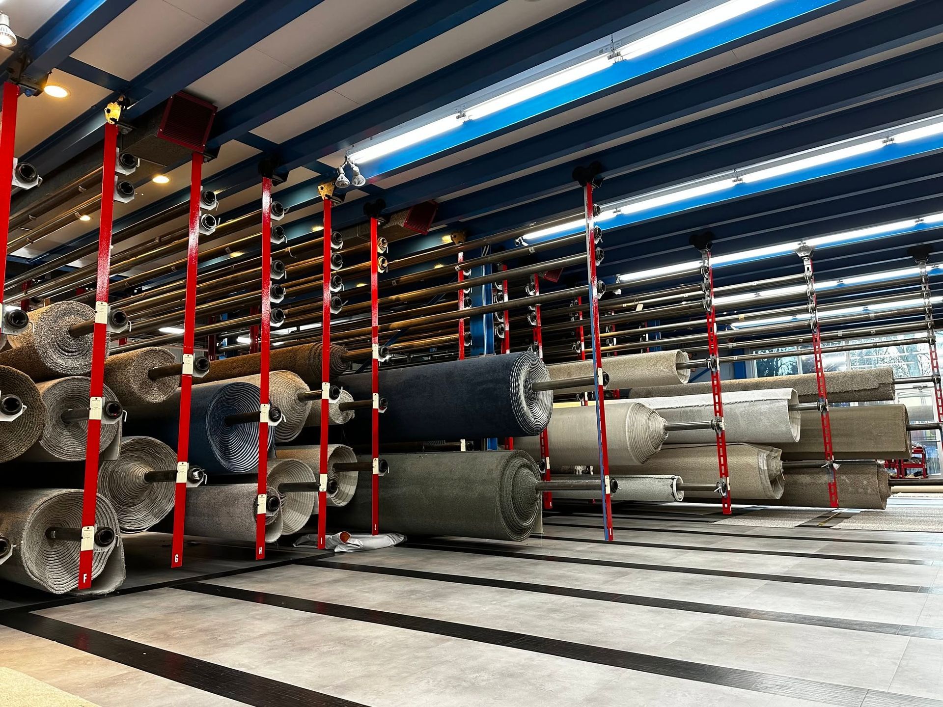 Rows of stacked carpet rolls in a warehouse, separated by red straps under bright blue ceiling lights.