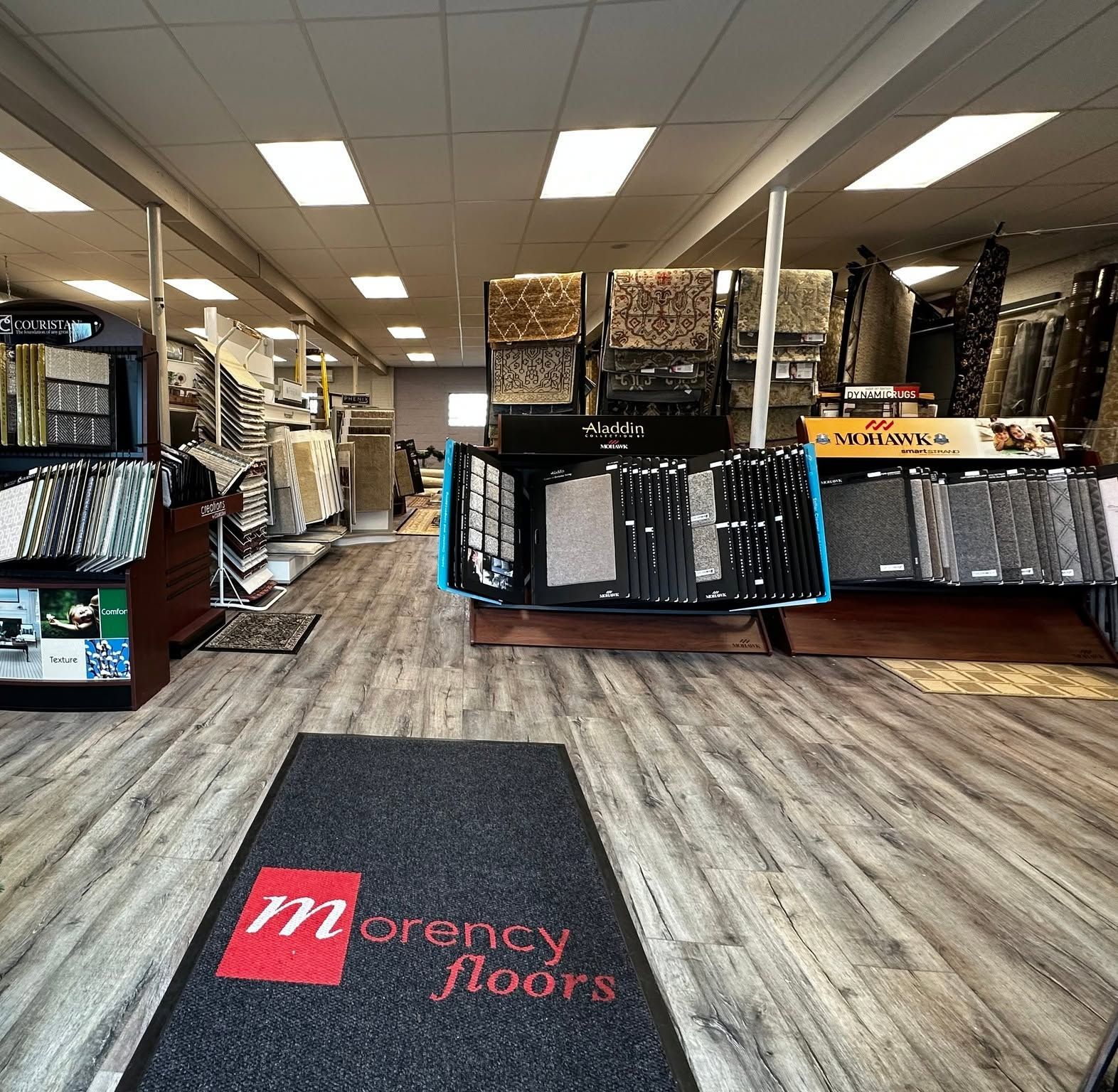 Flooring showroom with display racks, carpet samples, and a Morency Floors logo mat at the entrance