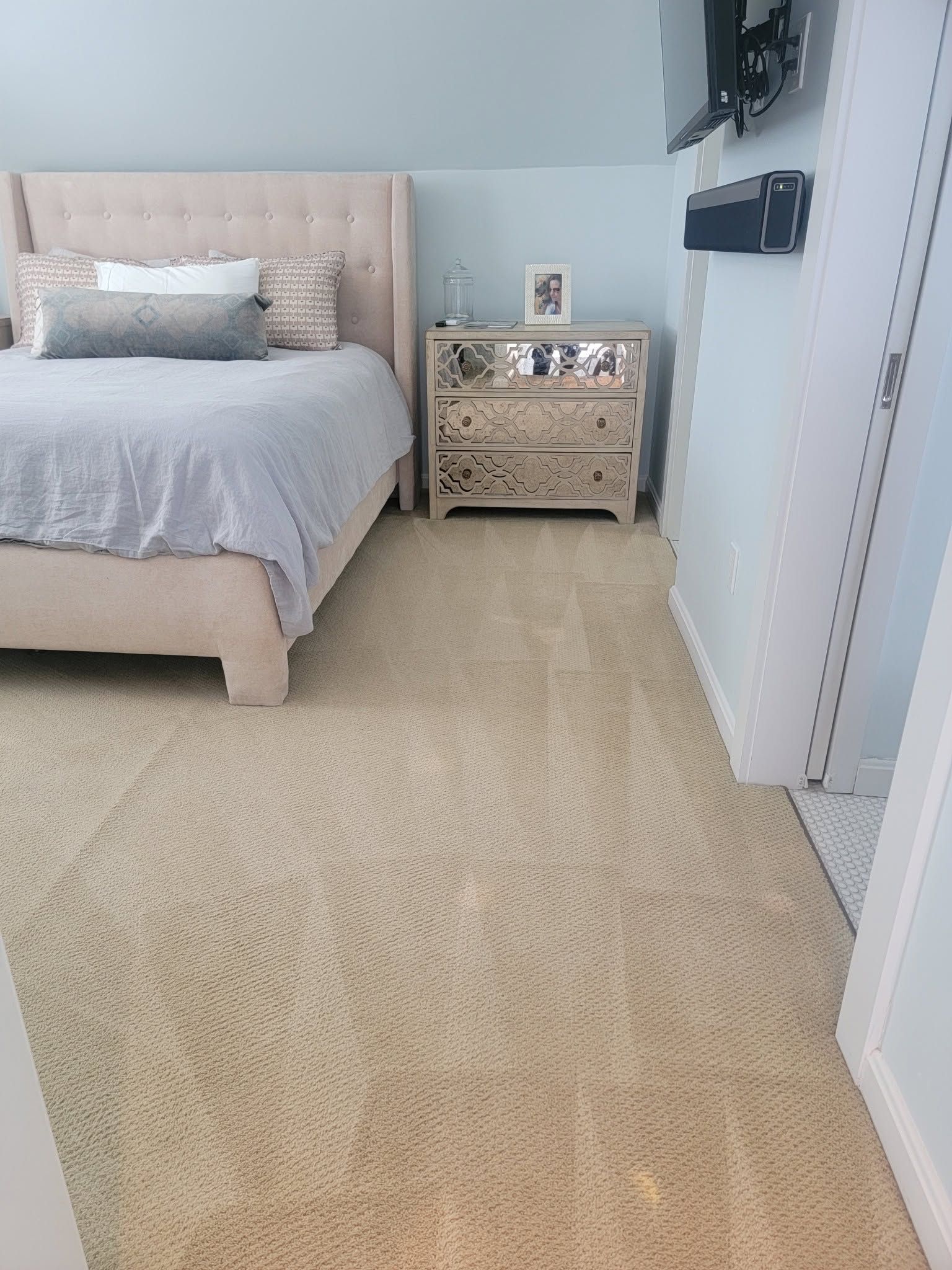 Bright bedroom with beige carpet, a bed, and a mirrored nightstand against light blue walls.