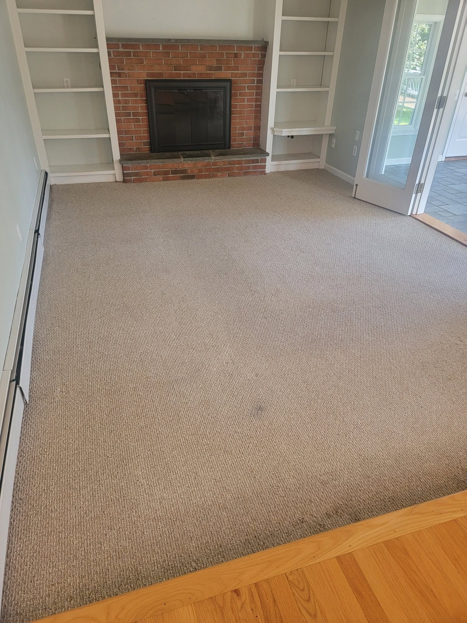 Empty living room with beige carpet, brick fireplace, built-in shelves, and sliding glass doors