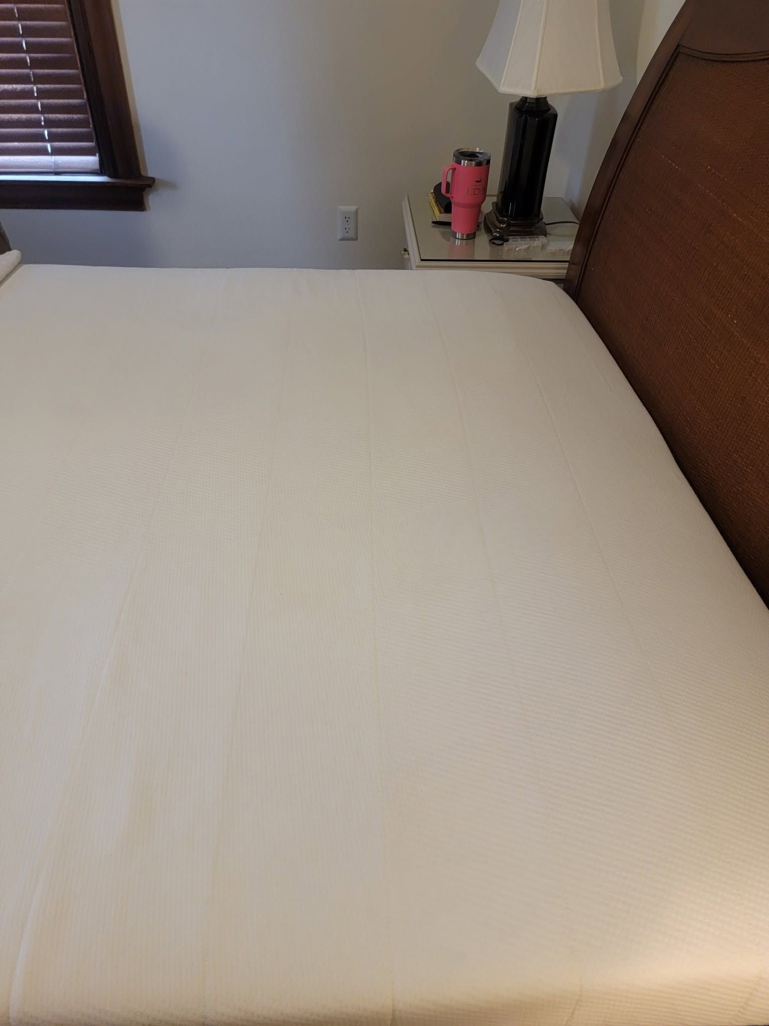 White bed in a bedroom beside a window and bedside table with lamp and pink tumbler.