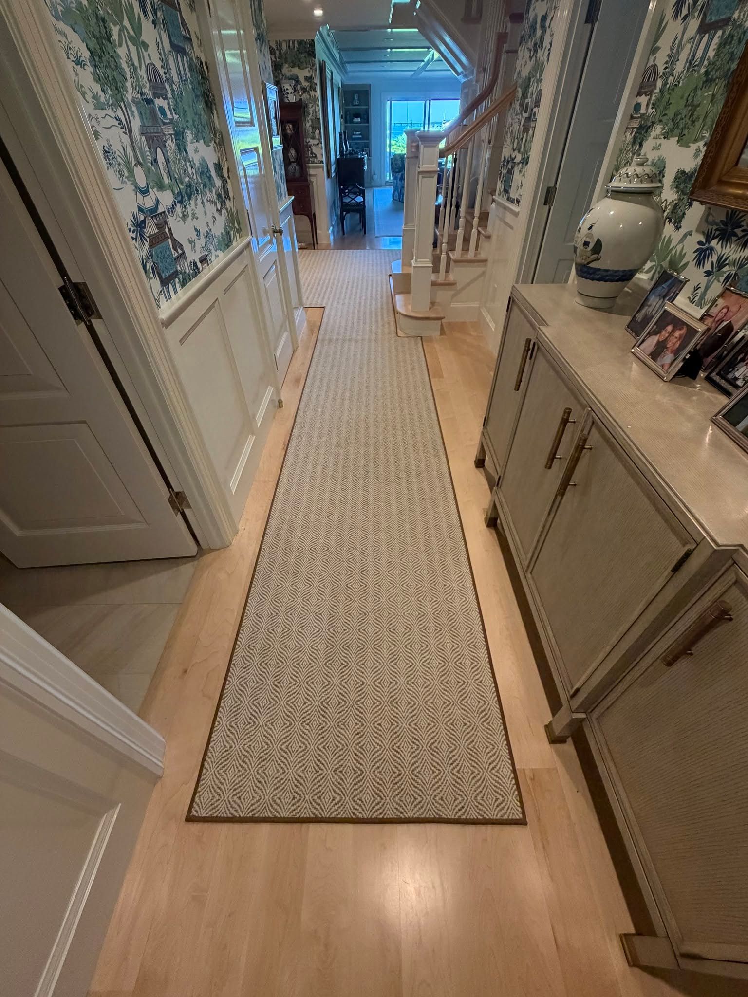 Narrow hallway with floral wallpaper, long runner rug, and cabinets on both sides, lit from the far end