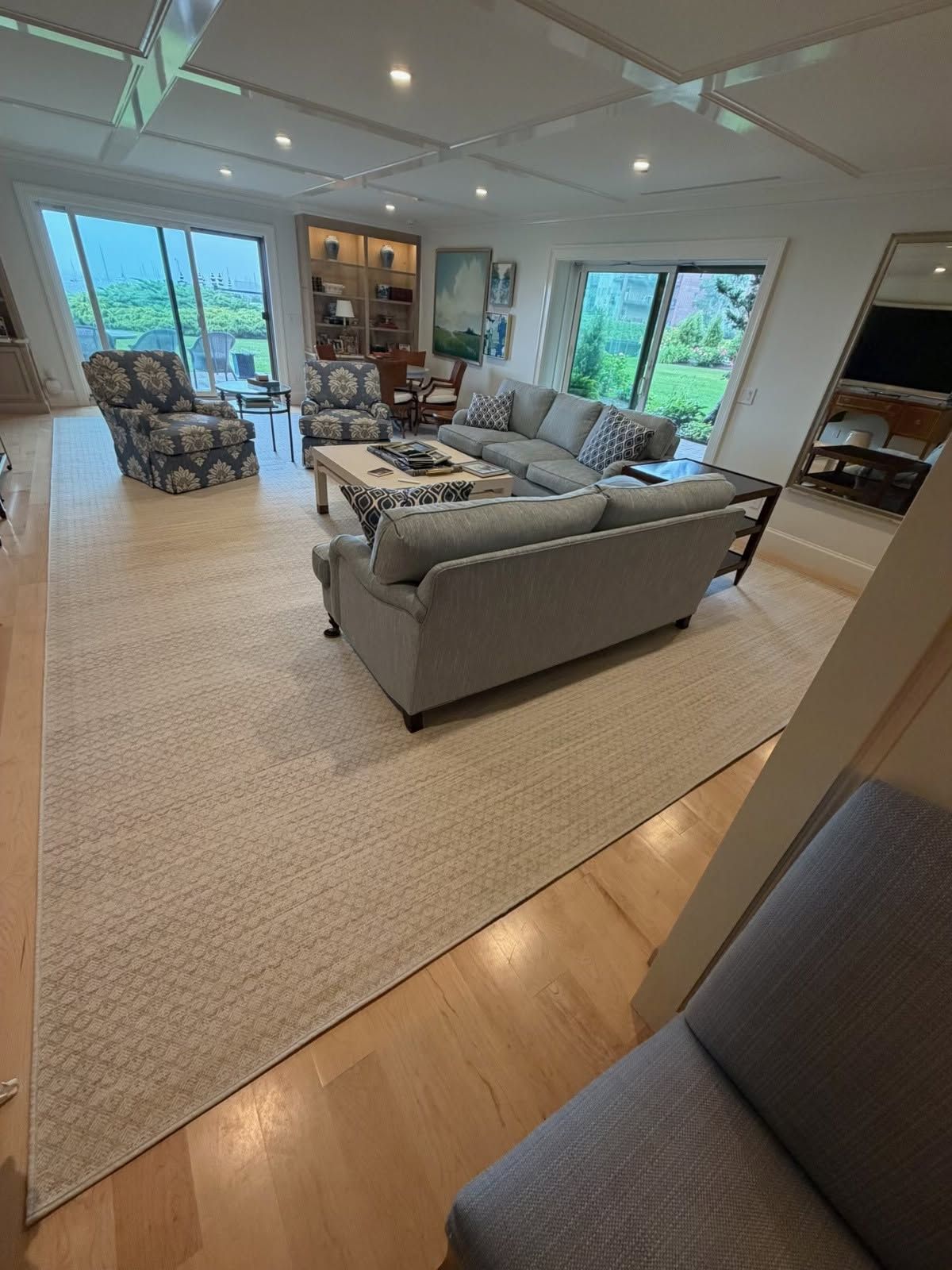 Modern living room with gray sectional, beige rug, wood floors, and large windows overlooking greenery