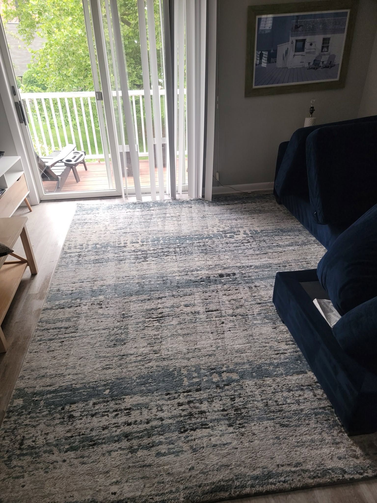 Living room with blue sofa and large patterned rug, open to a balcony through white vertical blinds.