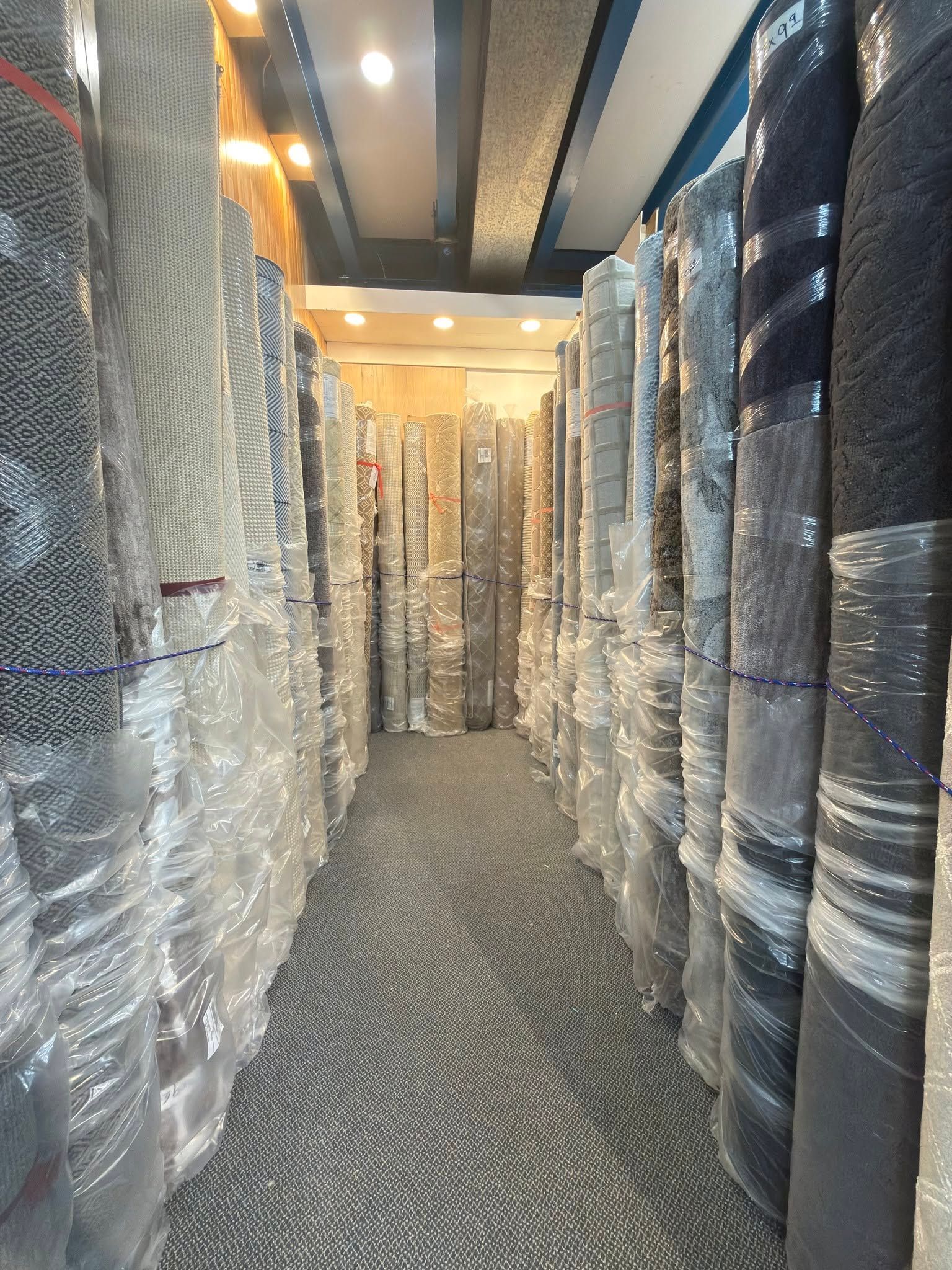 Narrow warehouse aisle lined with shrink-wrapped rolls or stacks of material under bright overhead lights