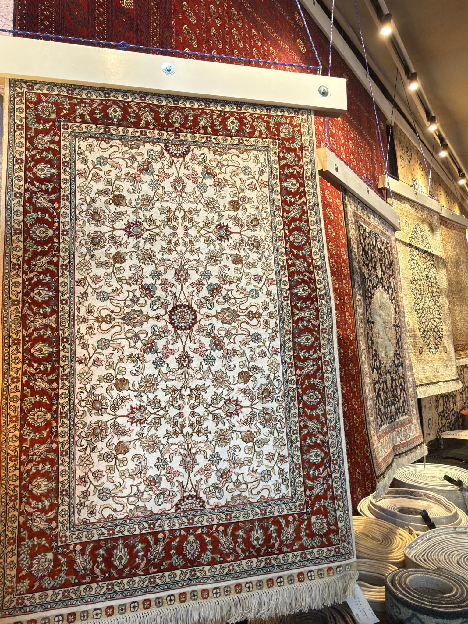Hanging rugs in a carpet shop, including a large white floral rug with red border.