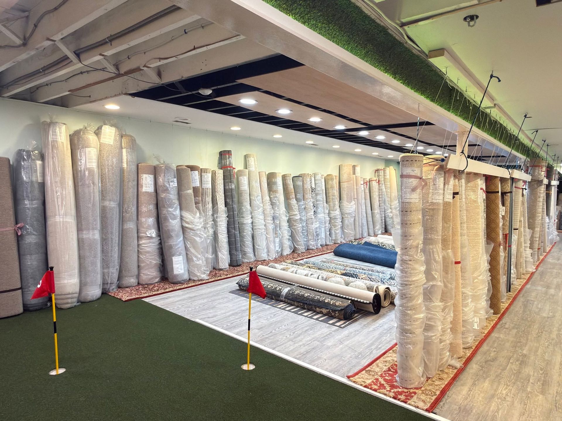 Carpet store aisle with rolled rugs and carpets displayed along the wall and floor samples in the center.