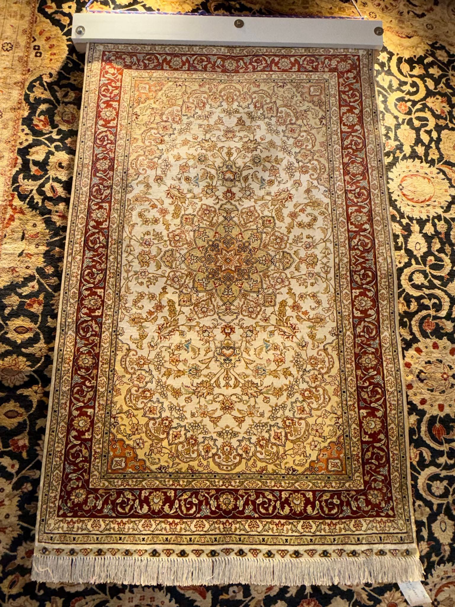Patterned beige and red prayer rug hanging on a wall display, with fringed bottom edge