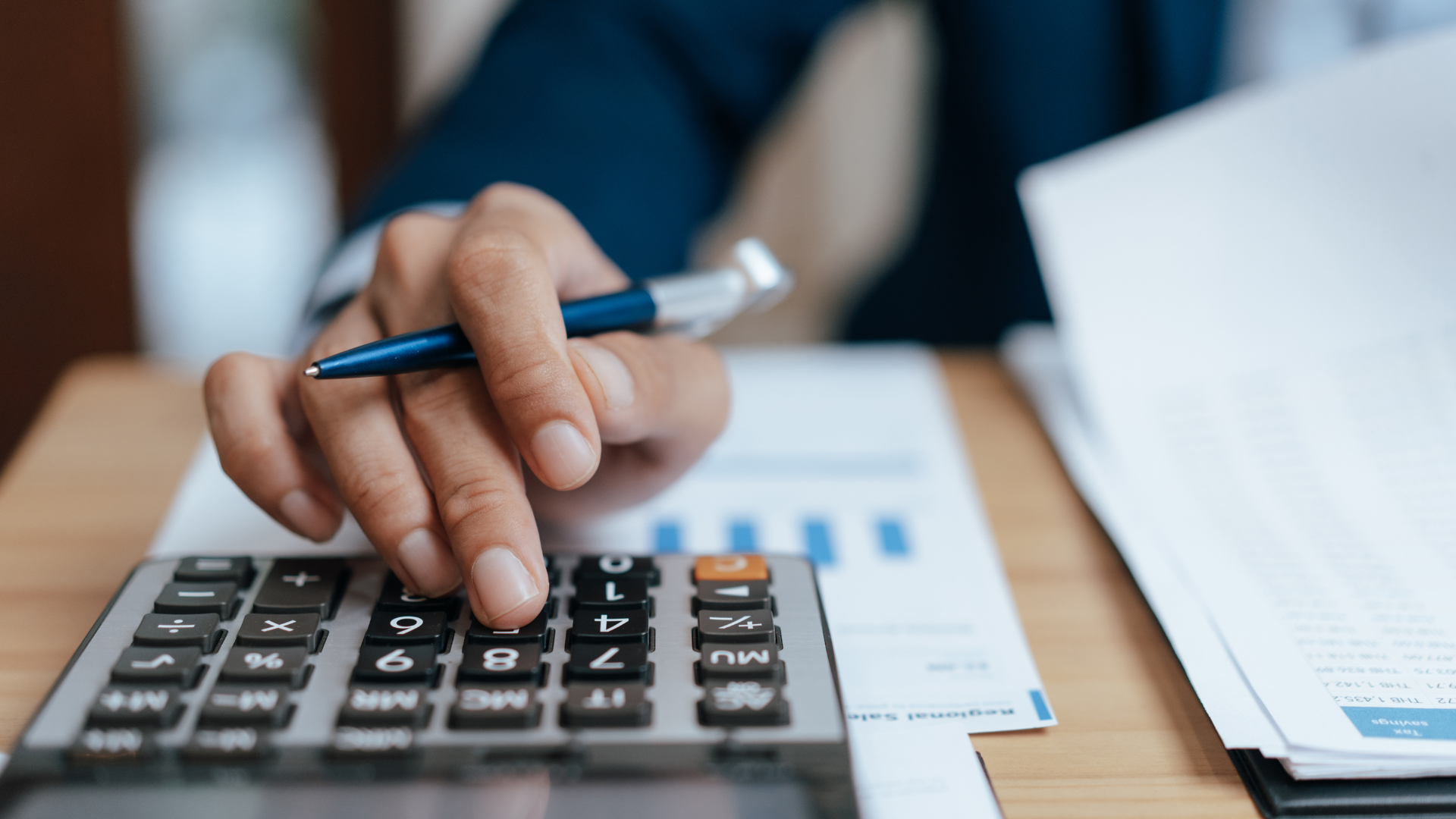 Hand counting on a calculator beside financial papers, with a pen on a desk