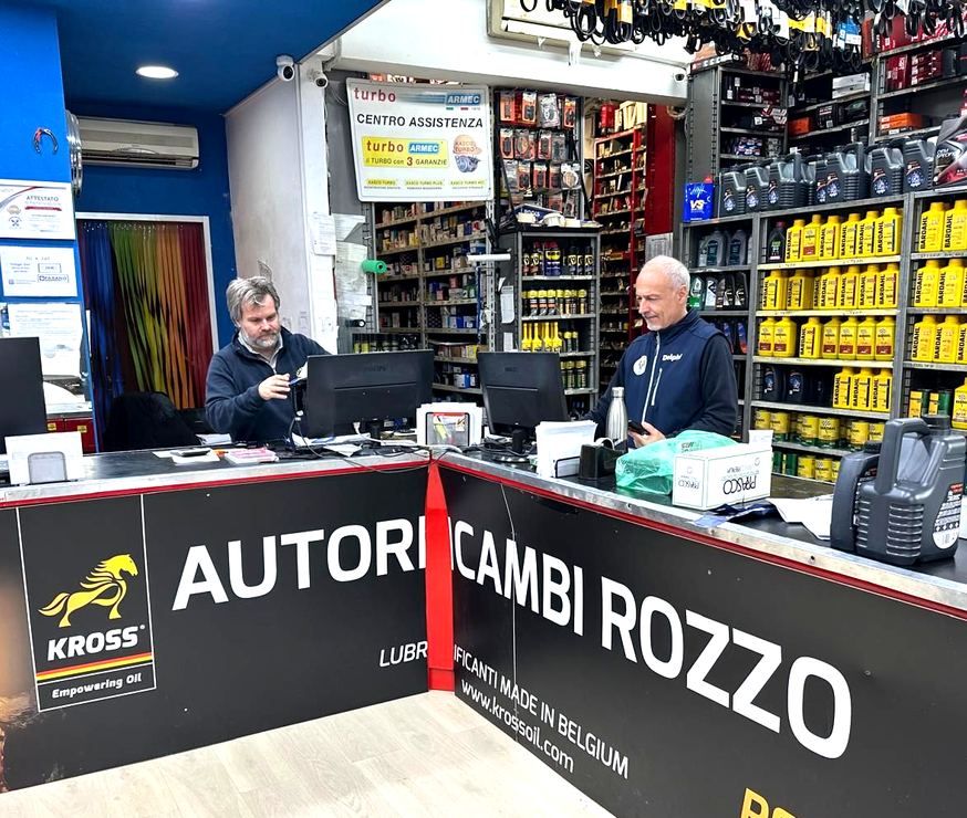 Tre uomini con le stesse camicie blu e le braccia incrociate posano davanti agli scaffali pieni di bottiglie di olio.