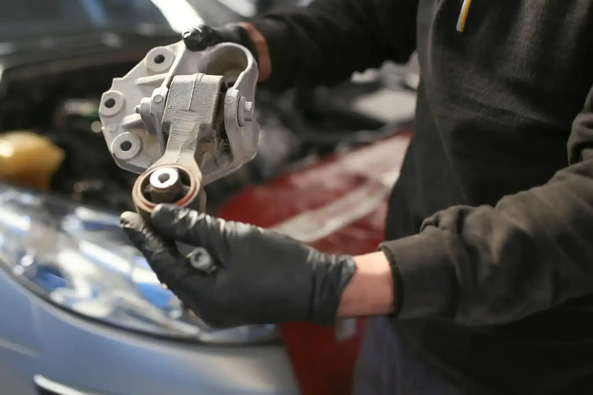 Meccanico che tiene in mano il supporto del motore di un'auto con guanti neri in un garage.