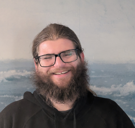 A smiling person with glasses, long hair, and a beard, wearing a black hoodie in front of a blue, wave-patterned background.