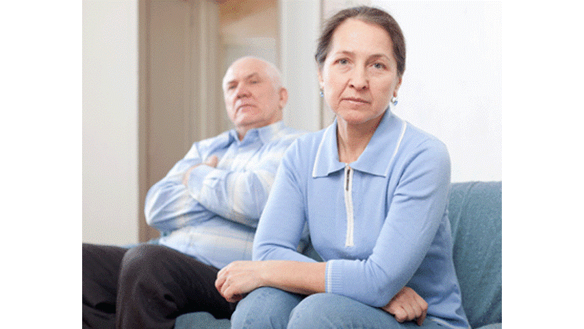 Woman in blue, looking serious, sits on a couch; man in background, arms crossed, seems displeased.