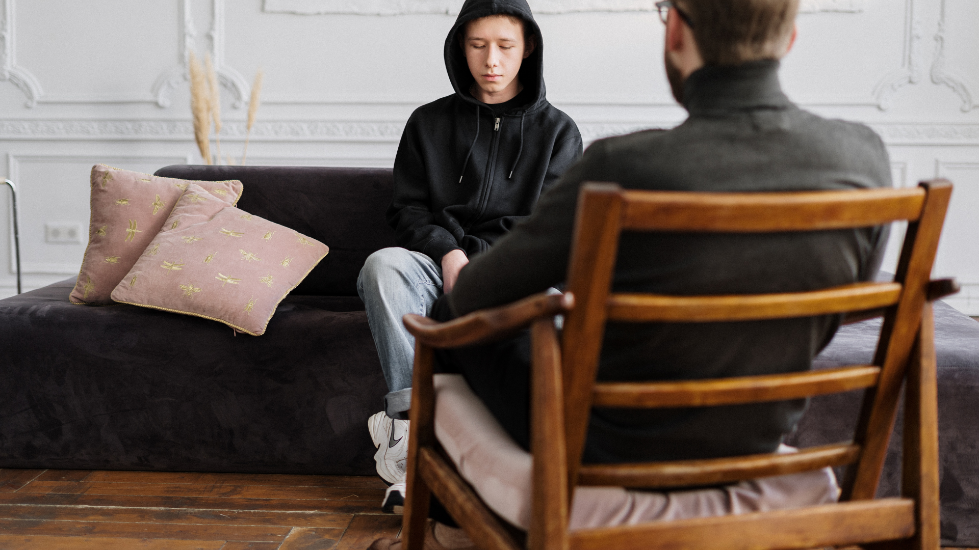 Two people in conversation across a living room, with a dark sofa and chair in the foreground.
