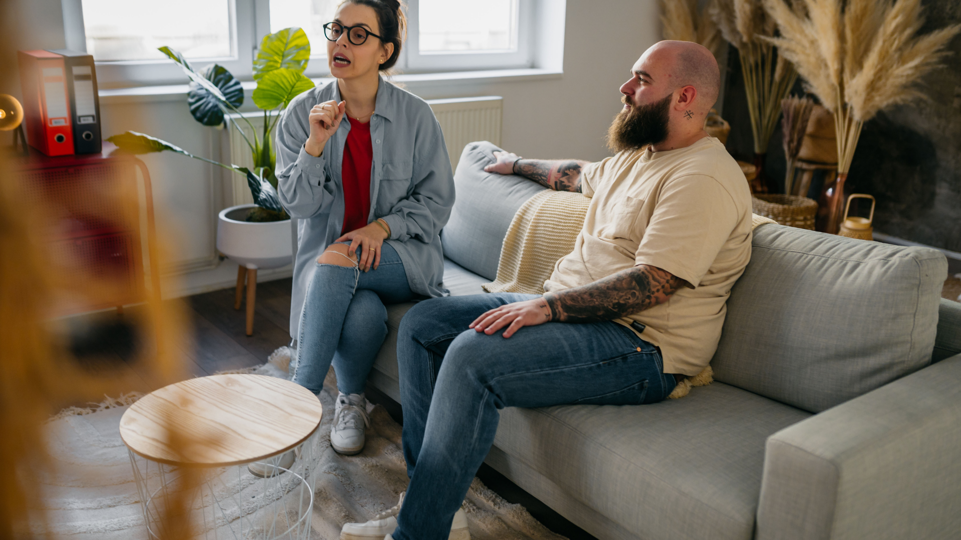 Two people talking on a gray couch in a bright living room with plants and a round wooden table.