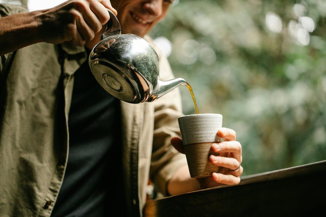 Person pouring coffee into a cup outdoors from a metal pot