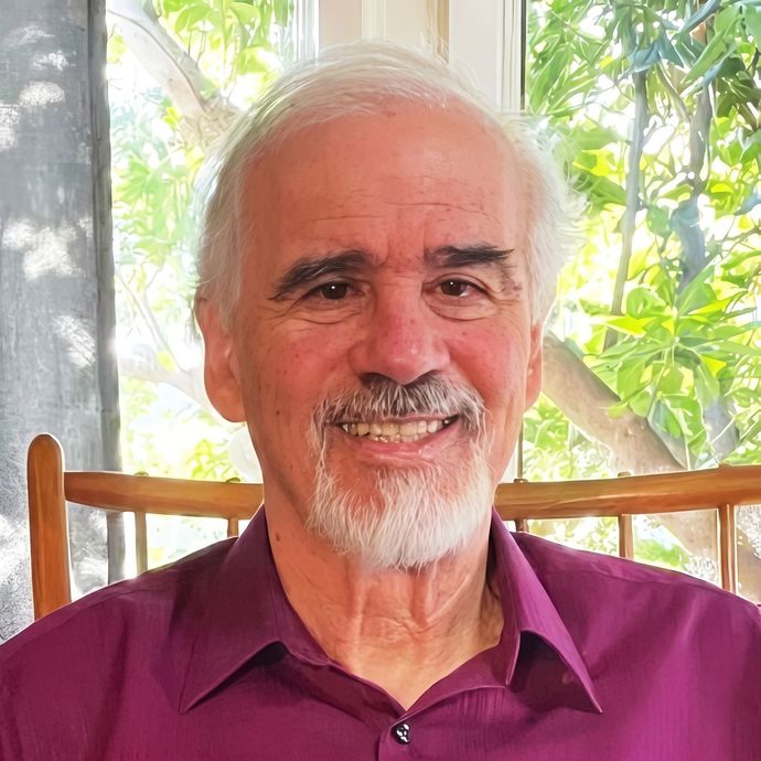 Smiling man with white hair and beard in a purple shirt, seated outdoors with green foliage behind him