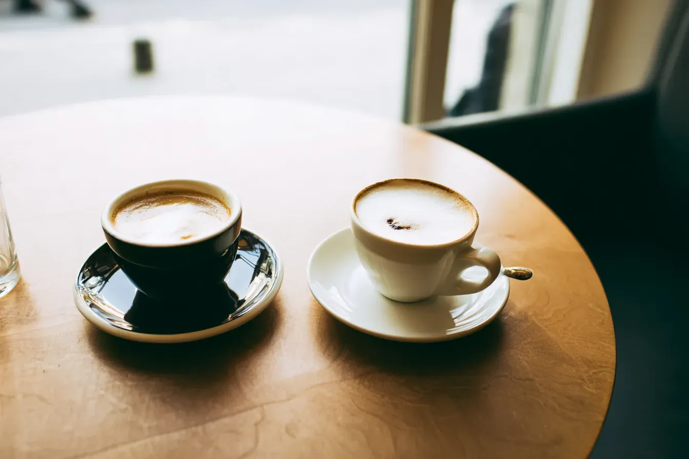 Two coffee cups on a wooden table, one in a black saucer and one in a white saucer near a window.