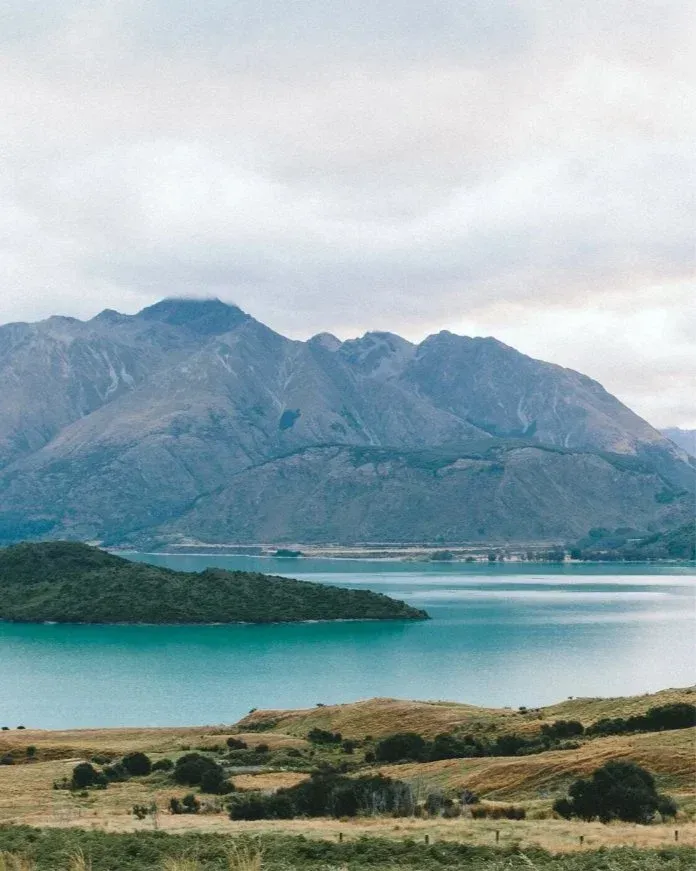 Turquoise lake below rugged mountains under a cloudy sky, with grassy hills in the foreground