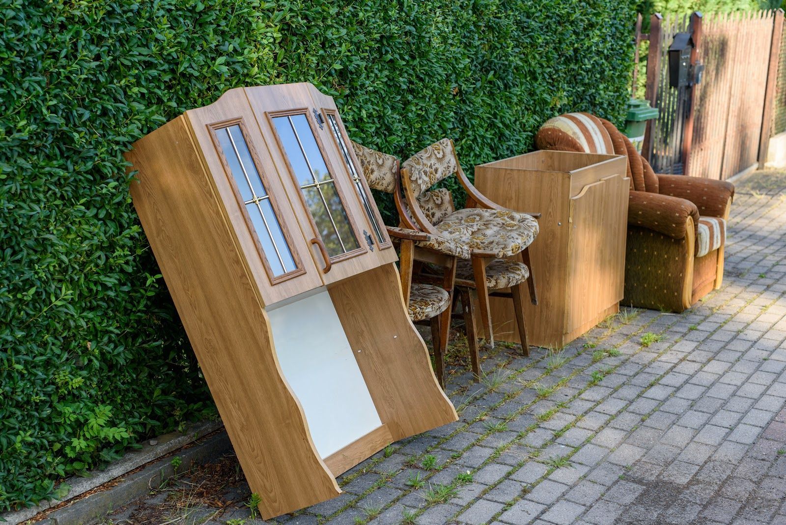 Outdoor display of wooden furniture and a small cabinet along a hedge-lined walkway