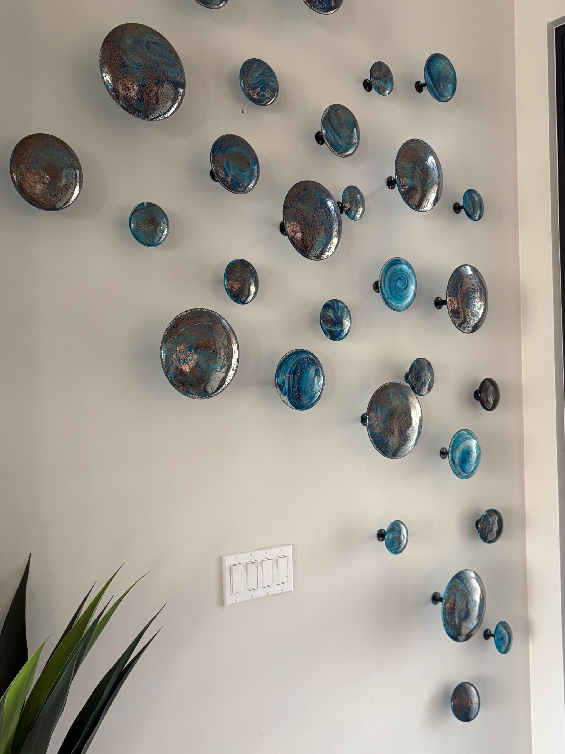 Wall art with iridescent, blue and silver disc-like shapes arranged diagonally on a light gray wall.
