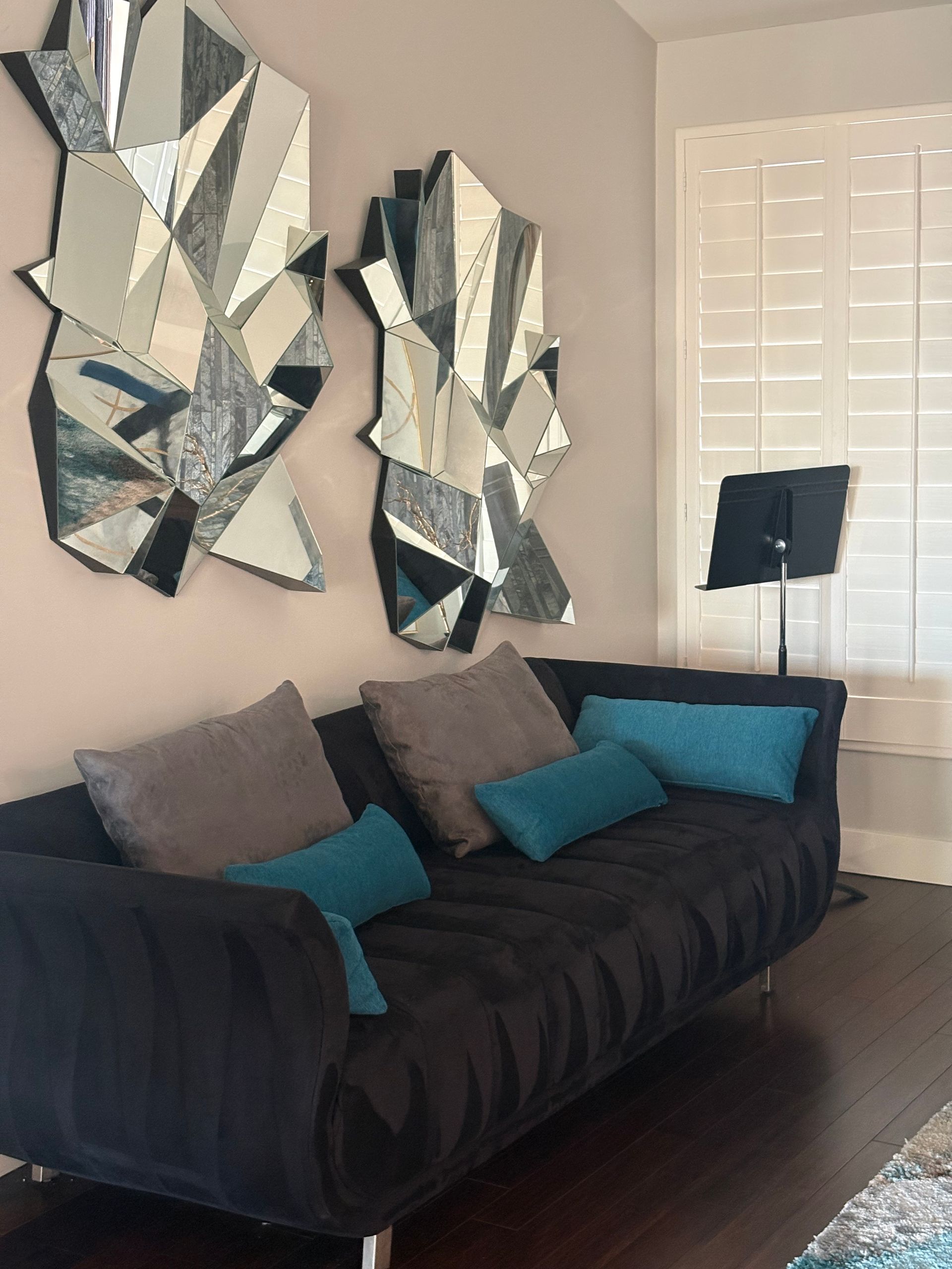 Black sofa with grey and turquoise pillows. Two geometric mirrors above. Shutters and music stand.