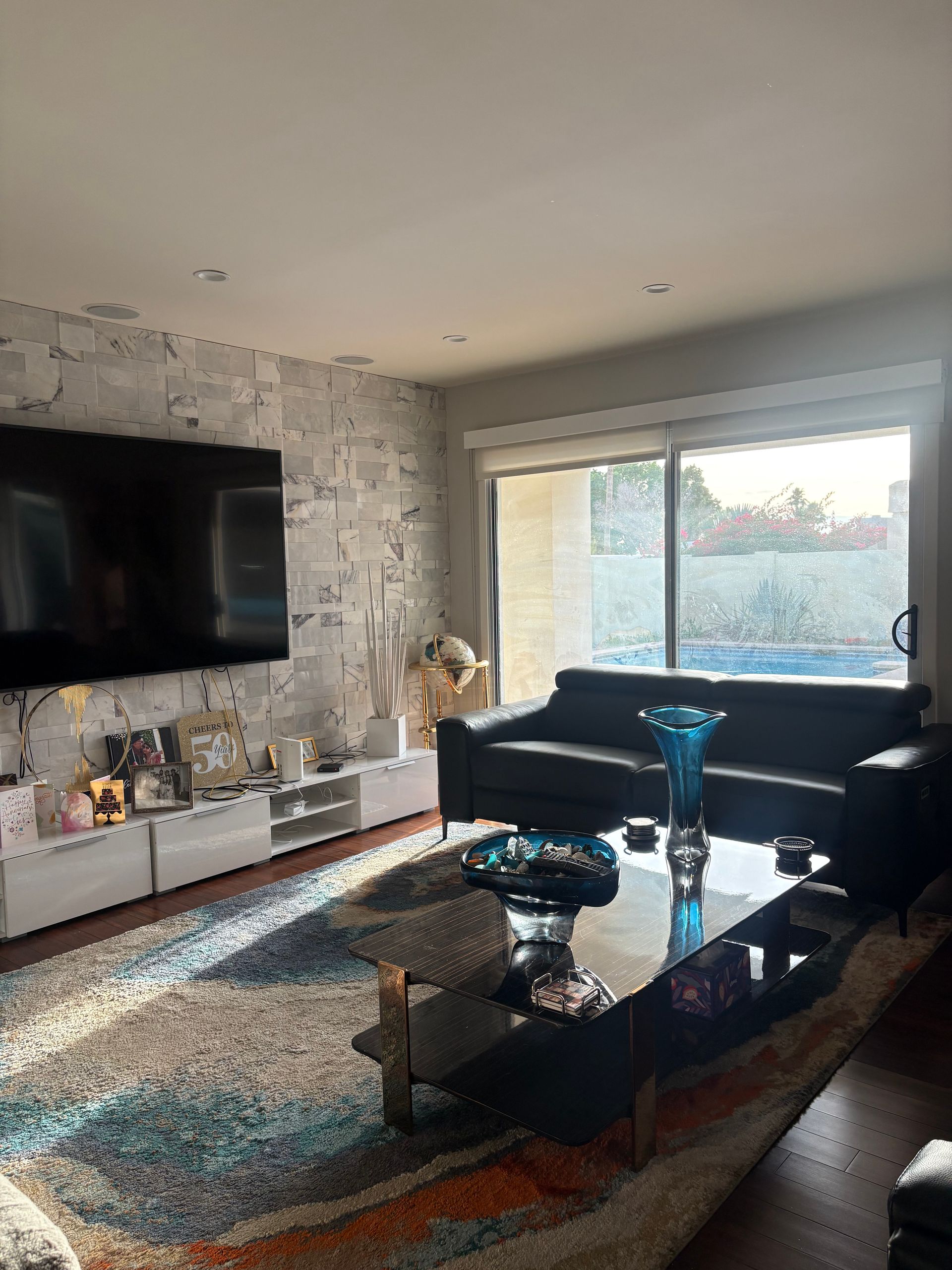 Living room with large TV, white wall, black sofa, glass table, and sunlight.