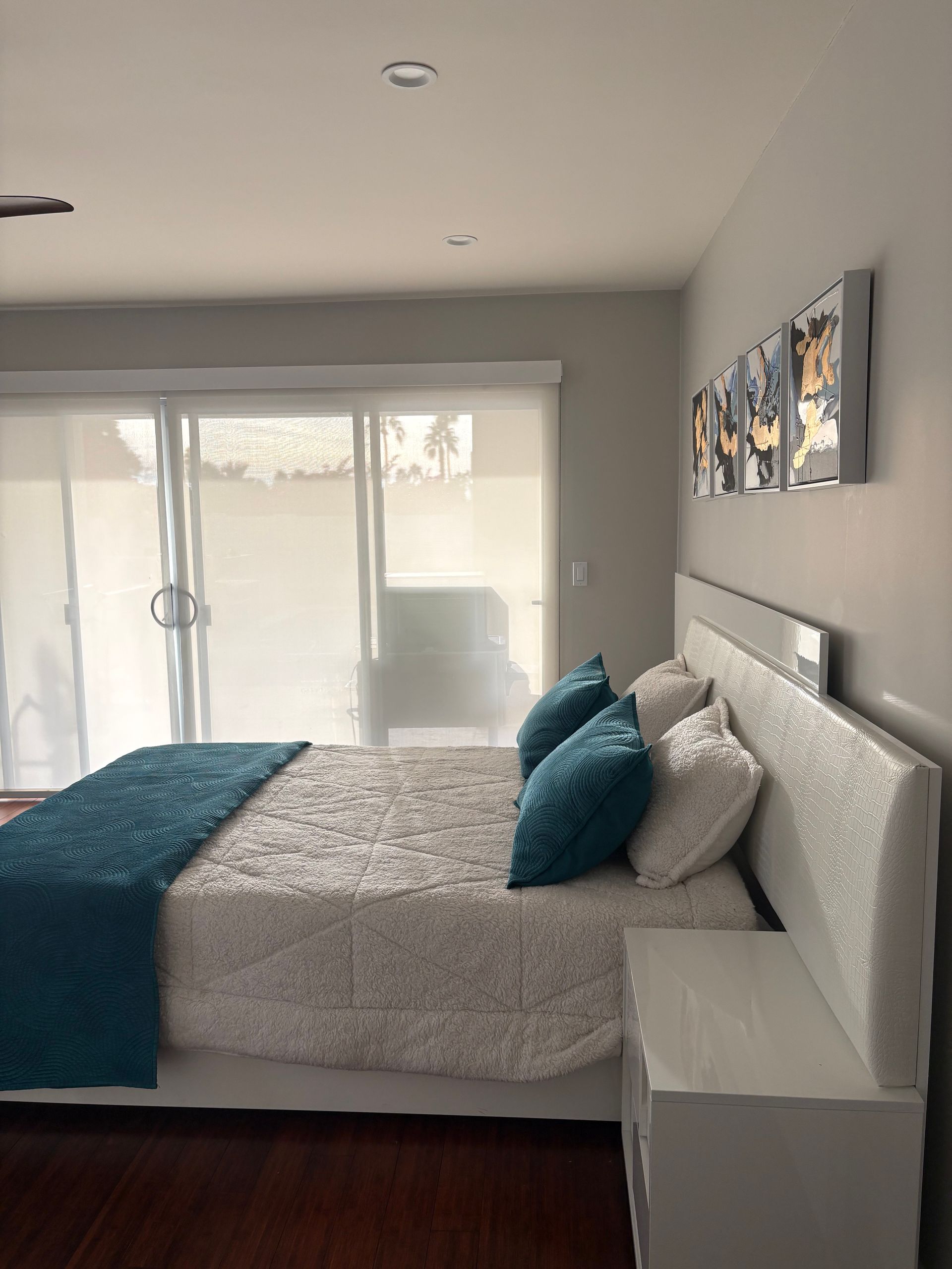 Bedroom with bed, blue throw, pillows, sliding glass door, white nightstand, art.