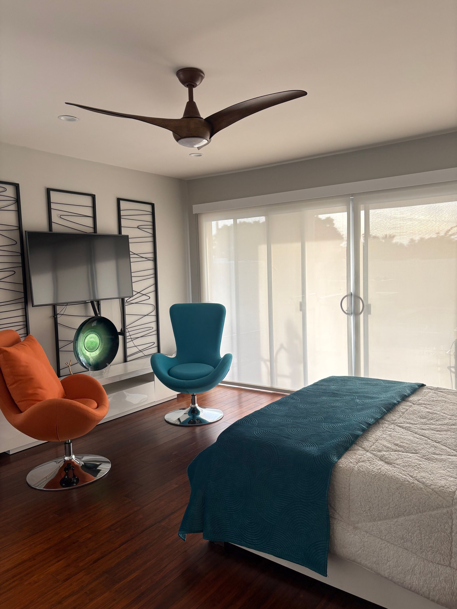 Bedroom with two swivel chairs, bed with teal throw, dark wood floor, and sliding glass doors. Brown ceiling fan.