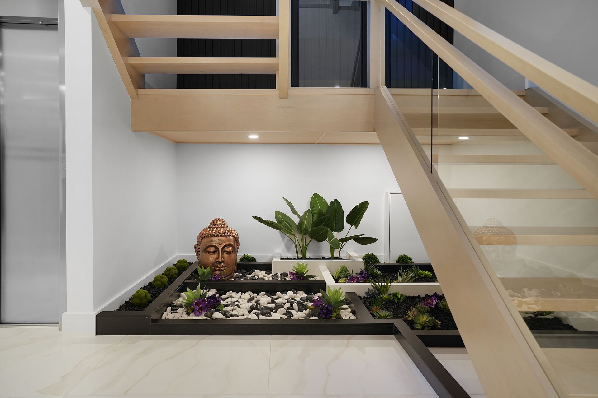 Indoor zen garden with a Buddha head statue, plants, and a staircase. The garden features dark wood trim, white and black stones, and greenery.
